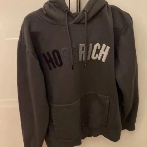 Mörkgrå hoodie från Hoodrich - Säljer en mörkgrå hoodie från Hoodrich med stor ficka framtill och justerbar huva. Texten 'Hoodrich' är broderad i olika nyanser av grått och svart på bröstet. Perfekt för en avslappnad stil.