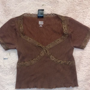 Brun ribbad topp från BDG - Säljer en brun ribbad topp från BDG med spetsdetaljer längs halsringningen och ärmsluten. Toppen har en omlott-design framtill och korta ärmar. Perfekt för en stilren look!