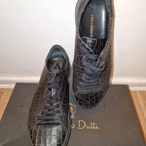 Svarta sneakers från Axel Arigato - Snygga svarta sneakers från Axel Arigato med ett unikt krokodilpräglat mönster. Skorna har klassisk snörning och en stilren design som passar perfekt för en trendig look. Sparsamt använda.