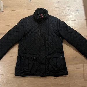 Svart quiltad jacka från Tommy Hilfiger - Snygg svart quiltad jacka från Tommy Hilfiger med dragkedja och knappar. Jackan har långa ärmar och praktiska fickor framtill. Perfekt för kyligare dagar med sin stilrena design.