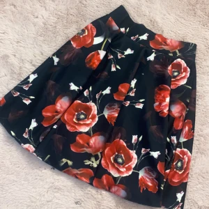 Svart kjol med rosa/röda blommor - Snygg svart kjol med stora rosa blommor och dragkedja i sidan. Kjolen har en vid passform och är perfekt för att ge en färgklick till din outfit. På bilderna ser kjolen lite orange röd ut men i verkligheten är den mer rosa🥰 Knappt använd