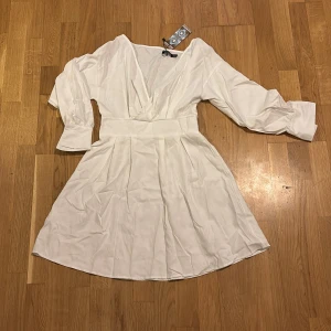 Vit klänning från Boohoo - Säljer en stilren vit klänning från Boohoo med v-ringad design och långa ärmar. Klänningen har en elastisk midja för en bekväm passform och är perfekt för sommarens alla tillfällen. Den är tillverkad i ett lätt och luftigt material. Nytt skick 