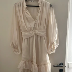 Vit kortklänning med volang från Bisou's Project - Superfin vit kortklänning från Bisou's Project med långa, luftiga ärmar och volangdetaljer nertill. Klänningen har v-ringning med knappar framtill och markerad midja. Perfekt för sommarens alla tillfällen!