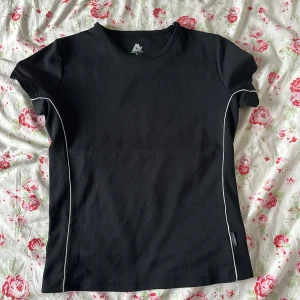 Svart träningst-shirt från Active Wear - Svart kortärmad träningst-shirt från Active Wear med vita kontrastsömmar längs sidorna. Tillverkad i ett lätt och ventilerande syntetmaterial som passar perfekt för aktiva dagar.