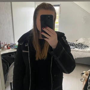 Svart teddyfodrad jacka med bälte - Säljer en svart jacka med teddyfoder och stora slag. Jackan har dragkedja, bälte i midjan och detaljer i skinnimitation. Perfekt för kyliga dagar och ger en cool look. Skinnet på fickorna är något slitna men inget man tänker på 