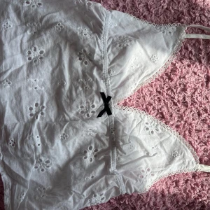 Vit broderad linne topp - Säljer en superfin vit linne topp med broderade blommor och en söt liten svart rosett framtill. Toppen är ärmlös och har smala axelband, perfekt för sommardagar. Den är i nyskick och passar både till vardags och fest!