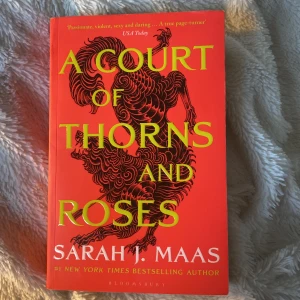 A Court of Thorns and Roses - Säljer boken 'A Court of Thorns and Roses' av Sarah J. Maas. Omslaget är rött med en svart drakillustration och gul text. Boken är på engelska och tillhör fantasygenren. Perfekt för dig som gillar spänning, magi och romantik.