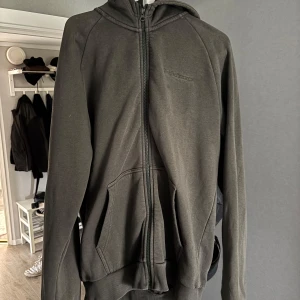 Mörkgrå hoodie med dragkedja från Peak Performance - Säljer en mörkgrå hoodie från Peak Performance med dragkedja framtill och två fickor. Tröjan har huva och ribbade muddar vid ärmslut och nederkant. Perfekt för en avslappnad stil.