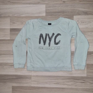Ljusblå sweatshirt med NYC-tryck från Ellos - Ljusblå sweatshirt från Ellos med stort NYC-tryck och texten 'New York City' på bröstet. Tröjan har rund halsringning och långa ärmar. Perfekt för en avslappnad och cool stil. Har en liten fläck på höger ärm.