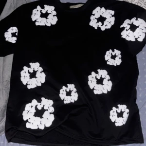 Svart t-shirt med vita blommönster från Denim Tears - Säljer en svart t-shirt från Denim Tears med flera stora vita blommönster tryckta över hela tröjan. T-shirten har en klassisk passform och är kortärmad, perfekt för dig som vill sticka ut med en unik design.