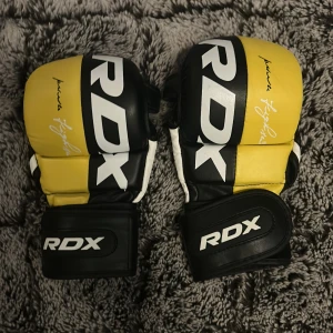 Svarta och gula mma handskar från RDX - Säljer ett par svarta och gula mma handskar från RDX med vit detalj och logga. Handskarna har öppen insida för bättre grepp och justerbar kardborreknäppning vid handleden. Perfekta för kampsport eller gymträning.