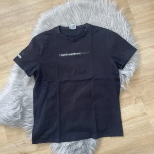 Svart t-shirt från Emporio Armani EA7 - Svart t-shirt från Emporio Armani EA7 med tryckt logga på bröstet och ärmen. Klassisk rund halsringning och normal passform. Perfekt för en stilren och sportig look.