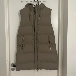 Beige lång dunväst med huva från Zara - Lång beige dunväst från Zara med huva och dragkedja framtill. Västen har två snedställda fickor med dragkedja och justerbara snören vid huvan. Perfekt för lager-på-lager-stil under kyliga dagar.