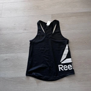 Svart träningslinne från Reebok - Säljer ett svart sportigt träningslinne från Reebok med stort vitt logotryck på sidan. Linnet har brottarrygg och är gjort i ett lätt och ventilerande material, perfekt för gym eller löpning.
