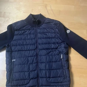 Svart pufferjacka från Moncler - Säljer en svart pufferjacka från Moncler med quiltad framsida och ärmar i stickad bomull. Jackan har dragkedja framtill och två fickor med dragkedja. Klassisk Moncler-logga på ärmen och hög krage för extra stilpoäng.