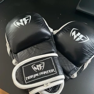 Svarta MMA-handskar från Nordic Fighter - Svarta MMA-handskar från Nordic Fighter med vita detaljer och öppna fingrar. Tillverkade i skinn med kardborreknäppning runt handleden för extra stöd. Perfekta för kampsportsträning och grappling.