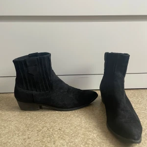 Svarta mocka boots - Säljer ett par svarta boots i mocka med låg klack och rund tå. Skorna har elastiska paneler på sidorna för enkel påtagning och en klassisk siluett som passar till många olika outfits.
