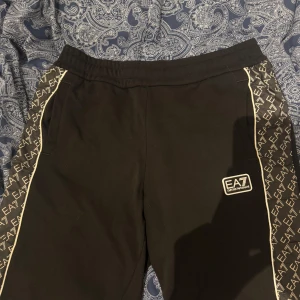 Svarta shorts från EA7 Emporio Armani - Svarta shorts från EA7 Emporio Armani med vita detaljer längs sidorna och tydliga loggor på benen. Shortsen har resår i midjan och en sportig look, perfekta för en avslappnad stil.