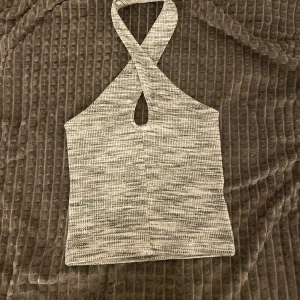 Gråmelerad ribbad halterneck topp - Säljer en gråmelerad ribbad halterneck topp med korsade axelband och en liten öppning framtill. Toppen är ärmlös och har en croppad passform, perfekt för en trendig look.