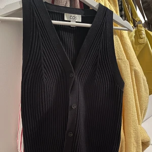 Svart ribbad väst från COS - Säljer en svart ribbad väst från COS med v-ringning och knappar framtill. Materialet är mjukt och stretchigt för en bekväm passform, perfekt till fest o middag! I stort sett oanvänd.