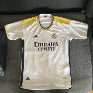 Real Madrid hemmatröja 2023/24 - Säljer en vit Real Madrid hemmatröja från Adidas med gula detaljer på axlarna och mörkblå inslag. Tröjan har klubbmärke och Adidas-logga på bröstet samt sponsortryck framtill. Klassisk v-ringning med gul och blå kant.