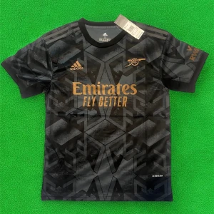 Arsenal svart fotbollströja från Adidas - Snygg svart Arsenal fotbollströja från Adidas med guldiga detaljer och loggor. Tröjan har korta ärmar och geometriskt mönster över hela tyget. Perfekt för dig som vill sticka ut på planen eller visa ditt stöd för laget.