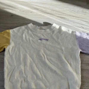 Vit ribbad tröja med färgade ärmar från H&M - Säljer en ribbad tröja från H&M i vitt med texten 'FREE FOREVER' broderad i lila på bröstet. Ena ärmen är gul och den andra lila, vilket ger en cool kontrast. Tröjan har rund hals och långa ärmar.
