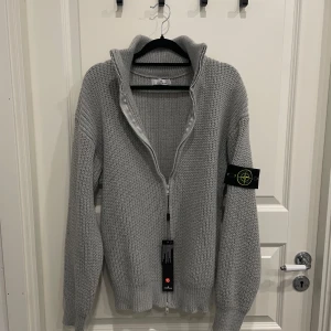 Stone Island Cardigan - Säljer en snygg ljusgrå stickad kofta från Stone Island med dragkedja framtill och hög krage. Klassisk patch på ärmen och ribbade muddar. Perfekt för lager-på-lager och stilren look.