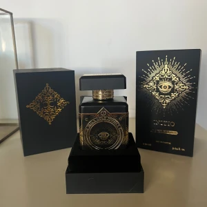 Initio Oud For Greatness Parfym - Lyxig parfymflaska från Initio med namnet Oud For Greatness. Flaskan är svart med guldiga detaljer och har ett mystiskt öga i centrum. Kommer i en elegant svart ask med guldfärgat mönster och text. Perfekt för dig som gillar exklusiva dofter.