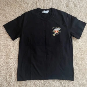 Svart t-shirt från Casablanca med tryck - Svart t-shirt från Casablanca med färgglatt drinkmotiv och texten 'CASABLANCA' på ryggen. Liten detalj på bröstet. Klassisk passform och rund halsringning. Perfekt för dig som gillar unika prints. Tröjan är i super bra skick bortsett från lite tryck som släppt, hör av dig om du har frågor.