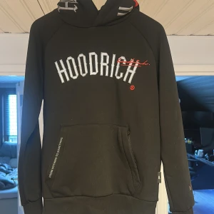 Svart hoodie från Hoodrich - Säljer en svart hoodie från Hoodrich med vit broderad logga framtill och tryck på ryggen. Tröjan har huva, känguruficka och ribbade muddar vid ärmslut och nederkant. Perfekt för en avslappnad streetwear-look.