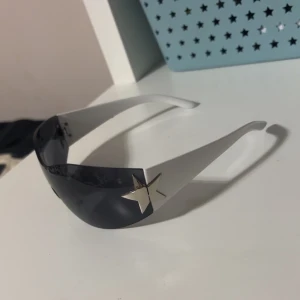 y2k Oversized svarta solglasögon med stjärndetaljer - Säljer ett par futuristiska oversized solglasögon med svarta glas och silvriga stjärnor på sidorna. Glasögonen har en sköldformad design utan synlig båge och breda skalmar i vitt. Perfekt statement piece för dig som vill sticka ut.