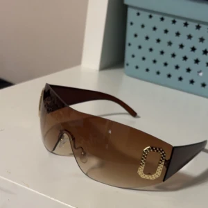 Bruna oversized y2k solglasögon  - Stora, bruna solglasögon med tonade linser och breda skalmar. På sidorna finns dekorativa guldfärgade detaljer med glittrande stenar. Perfekt accessoar för en trendig look.