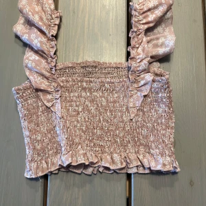 Rosa blommig croppad topp från Shein - Supersöt rosa croppad topp från Shein med blommigt mönster och volangdetaljer. Toppen har breda axelband med volanger och smockad resår över hela plagget för en bekväm passform. Perfekt för varma dagar!