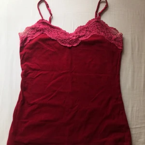 Söt röd linne med spets  - Säljer en röd linne från Lindex med tunna axelband och vacker spetsdetalj upptill. Toppen har blekfläckar under armhålan.