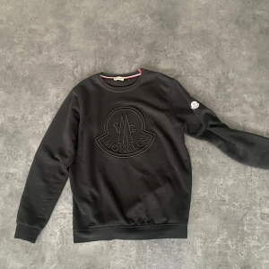 Svart sweatshirt från Moncler - Säljer en svart sweatshirt från Moncler med stort broderat Moncler-logo på bröstet och en patch på ärmen. Tröjan har rund halsringning och långa ärmar. Perfekt för en stilren och avslappnad look. Snygg som tusan 