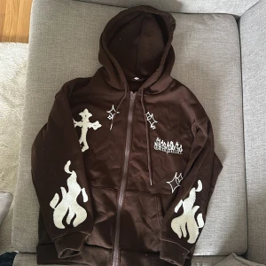 Brun hoodie med vita tryck - Cool brun hoodie med dragkedja och huva. Har vita tryck med eldflammor, kors och stjärnor på framsidan och ärmarna. Perfekt för dig som vill sticka ut med en unik streetstyle.