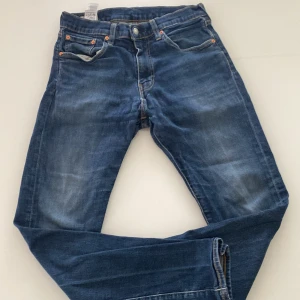 Levis blå jeans - Klassiska blå Levis 501 jeans med raka ben och fem fickor. Jeansen har knappgylf och tydliga Levis-detaljer som läderpatch bak och röd etikett på bakfickan. Perfekta för en tidlös denimlook.