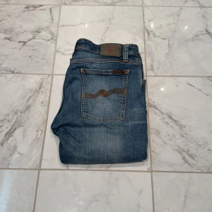 Blå jeans från Nudie Jeans - Säljer ett par klassiska blå jeans från Nudie Jeans med orange sömmar och snygga detaljer på bakfickorna. Jeansen har normal passform och raka ben, perfekt till vardags. Ikonisk Nudie-lapp i midjan. Nypris 1800kr mitt pris 499kr💯