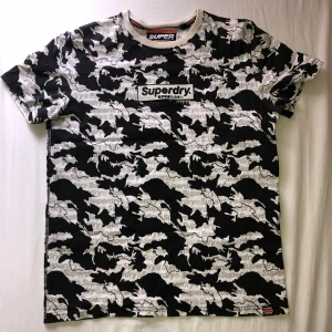 Svartvit camo t-shirt från Superdry - Säljer en svartvit t-shirt från Superdry med camouflagemönster och tydlig logga på bröstet. (“T” lifter lite som man ser i bild 2, säkert går att limma)