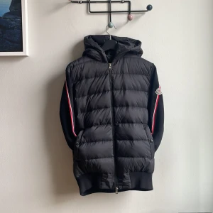 Svart cardigan jacka med luva från Moncler - svart cardigan från Moncler med huva och dragkedja framtill. Jackan har quiltad framsida, ribbade muddar och Moncler-logga på ärmen. Röda och vita ränder längs ärmarna. Perfekt för våren och hösten. Den har några små omärkbara fläckar på vänstra sidan om man tvättar så borde det försvinna. Jag är 187 cm på bilden