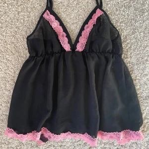 Babydoll linne - Söt svart linnetopp med rosa spetsdetaljer längs v-ringningen och nederkanten. Tunn och luftig med smala axelband och lätt utställd passform. I mesch tyg. Elastiskt band under bysten.
