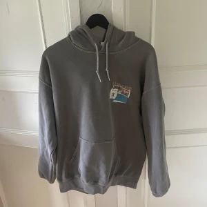 Grå hoodie med japanskt tryck|Urban outfiters  - Säljer en grå hoodie med stor färgglad print på ryggen och mindre tryck på bröstet. Tröjan har huva med dragsko och en klassisk magficka. Perfekt för dig som gillar streetwear och unika motiv.