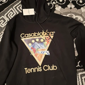 Casablanca tröja (svart)  -   Säljer en exklusiv Casablanca Tennis Club-huvtröja i svart. Denna hoodie är dekorerad med en unik grafisk print som inkluderar en triangel, pyramid, himlakroppar, tropiska växter och en tennisbana. Den har också en liten Casablanca Tennis Club-tagg på insidan av huvan. Tröjan är i utmärkt skick, knappt använd och perfekt för både vardagsbruk och sportiga tillfällen. Storlek: [M].  
