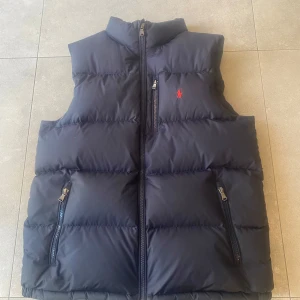 Marin blå dunväst från Polo Ralph Lauren - Säljer en marin blå dunväst från Polo Ralph Lauren med dragkedja framtill och två sidofickor med dragkedja. Västen har en klassisk passform och en liten röd broderad logga på bröstet. Perfekt för lager-på-lager under kyliga dagar. Passar nog s/m mått på bild 5. Pris kan diskuteras.