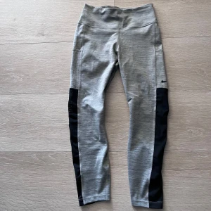 Grå träningstights från Nike - Snygga grå träningstights från Nike med svarta paneler längs benen. Hög midja och tight passform, perfekta för gymmet eller löpning. Diskret Nike-logga på låret. Bekvämt och stretchigt material.
