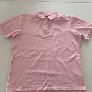 Ljusrosa pikétröja från Fila - Säljer en ljusrosa pikétröja från Fila med klassisk krage och tre knappar framtill. Tröjan har korta ärmar och diskret Fila-logga broderad på bröstet. Perfekt för en sportig och avslappnad stil.