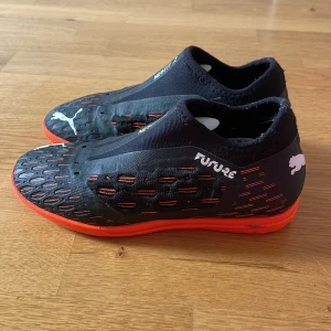 Puma Future fotbollsskor i svart och orange - Säljer ett par svarta och orangea Puma Future fotbollsskor med låg profil och mönstrad ovansida. Skorna har en tight passform och är tillverkade i syntetmaterial. Perfekta för dig som vill ha bra grepp och kontroll på planen. Pumas net-fit är som riktigt tyg fast mer slitstarkt! Super snygga och jättebra!⚽️