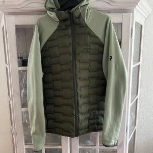 Grön hybridjacka med huva från Peak Performance - Säljer en grön hybridjacka från Peak Performance med quiltad front och mjuka ärmar. Jackan har huva och dragkedja hela vägen. Perfekt för lager-på-lager och outdoor-aktiviteter.