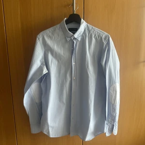 Ljusblå skjorta från Massimo Dutti - Snygg ljusblå skjorta från Massimo Dutti med klassisk krage och vita knappar. Skjortan har lång ärm och förstärkta armbågslappar i vitt. Tillverkad i bomull för en skön känsla och stilren look. Använd en gång.